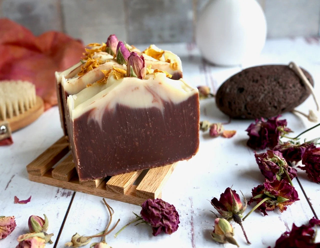 Bar of soap with decorative elements on a wooden stand, surrounded by dried flowers and a pumice stone scrubber.